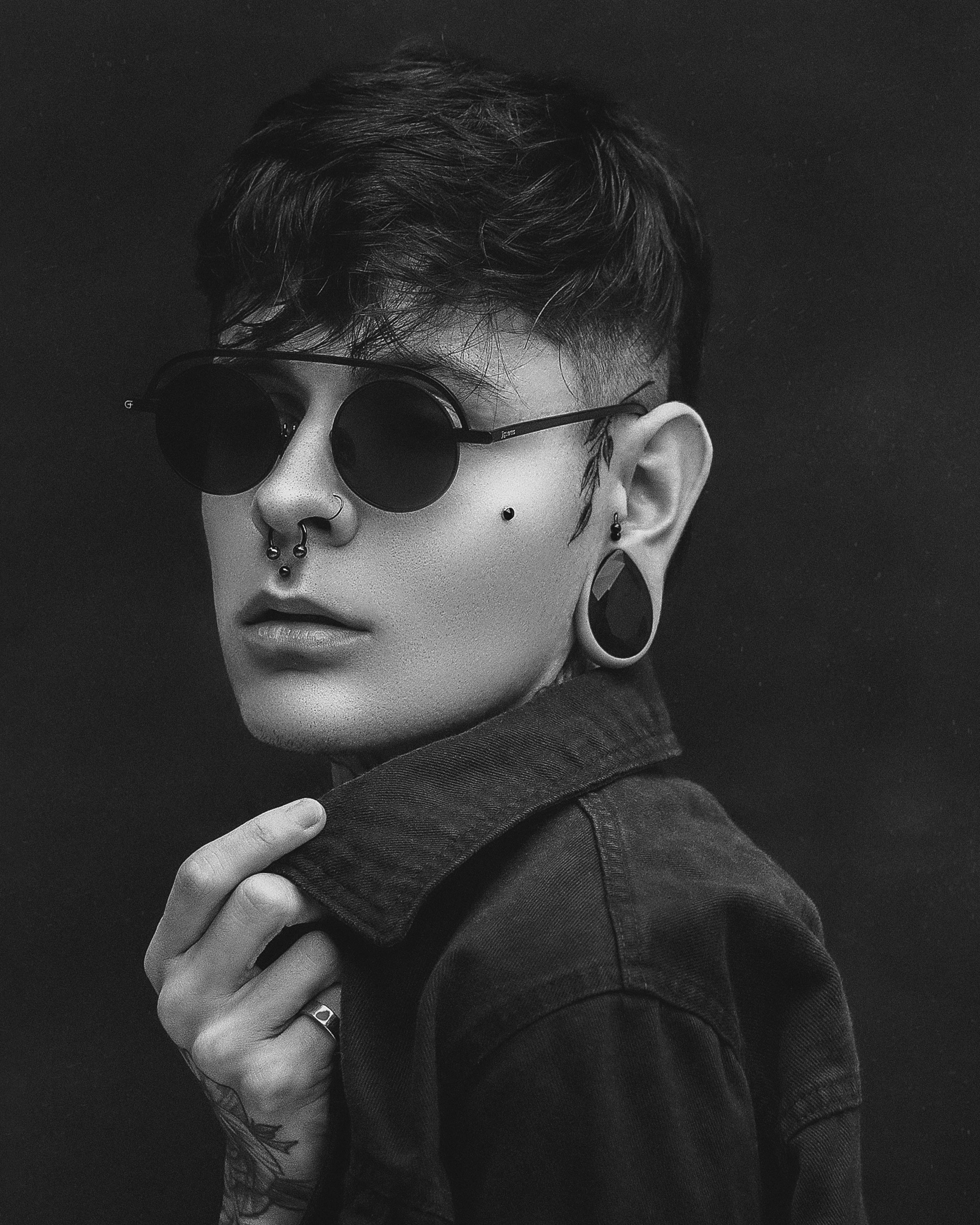 Stylish black and white portrait of a young adult wearing sunglasses and piercings.