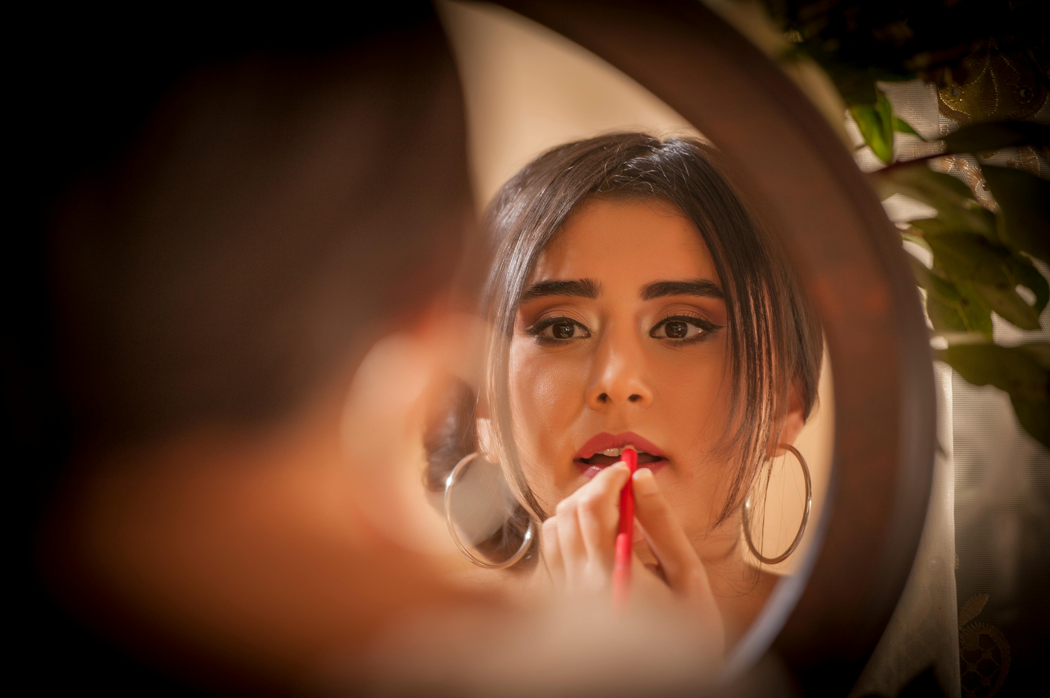 A woman applies red lipstick while looking into a mirror, reflecting a moment of elegance and beauty.