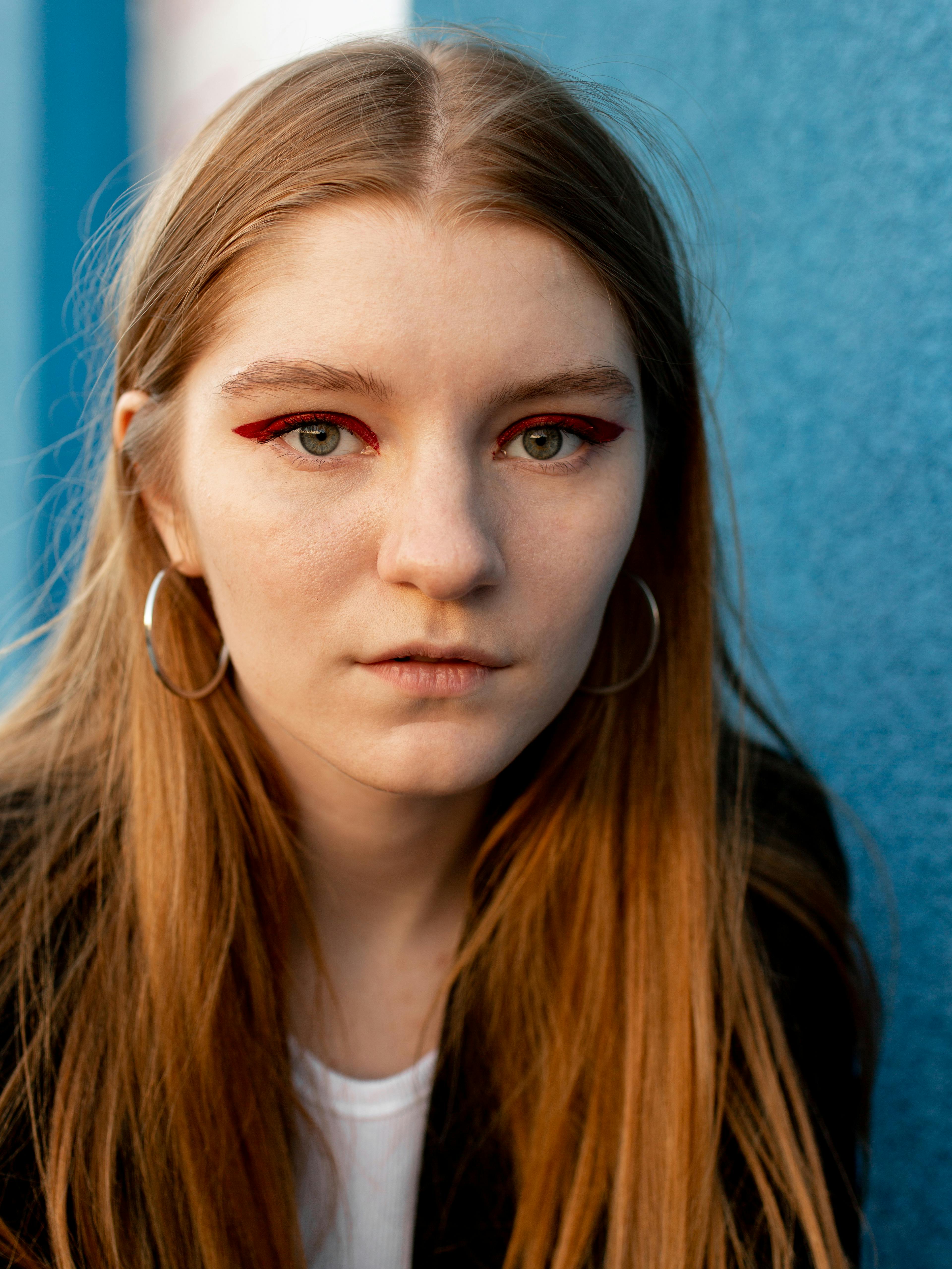 Close-up portrait of a young woman with bold red eye makeup and hoop earrings against a blue background.