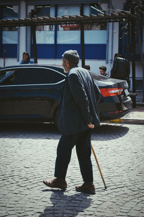 Free Senior man walking with a cane on a cobblestone street beside a black car. Stock Photo