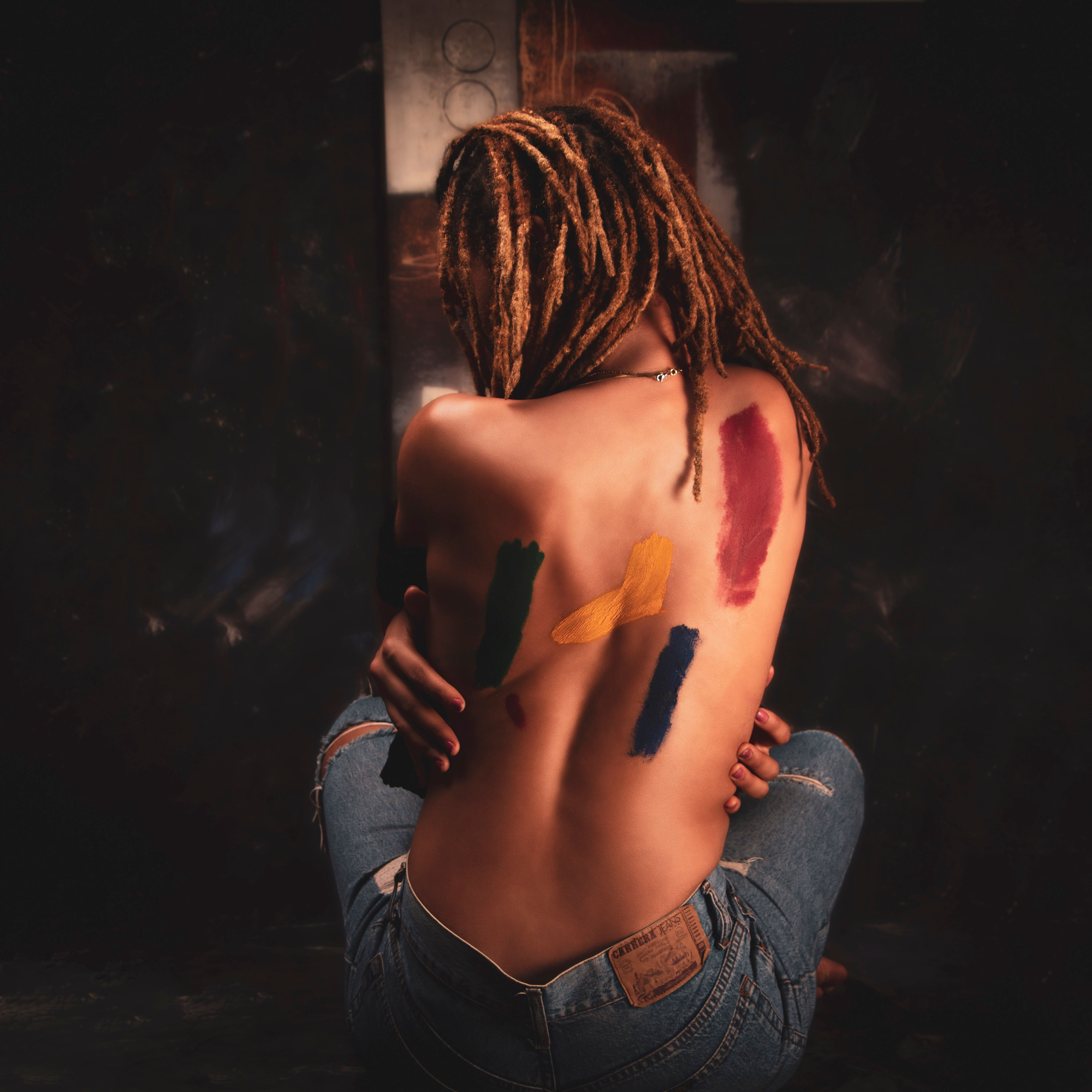 Back view of a person with painted strokes on their skin, dreadlocks, and wearing ripped jeans.