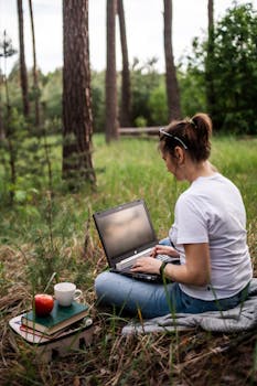 Young woman working remotely in a serene forest setting with laptop, books, and coffee.
