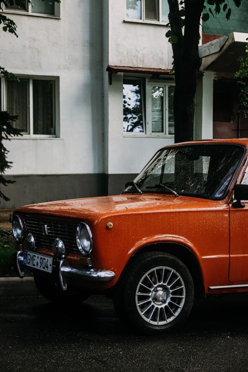 Free Retro orange car parked on a wet street near an urban building. Stock Photo