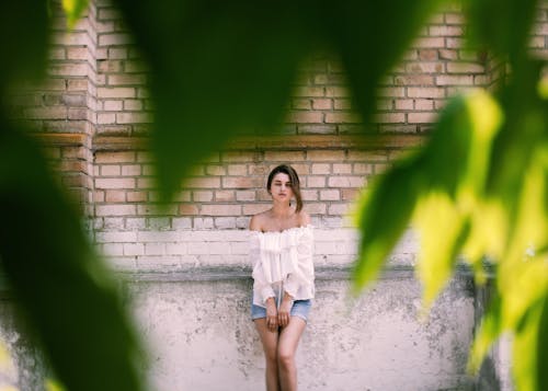 Free Woman in White Off Shoulder Shirt and Blue Denim Shorts Standing Beside Brick Wall Stock Photo