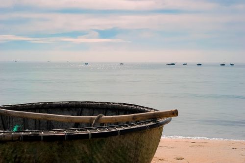 Free A rustic wooden rowboat rests on a peaceful beach with distant boats on the calm sea. Stock Photo