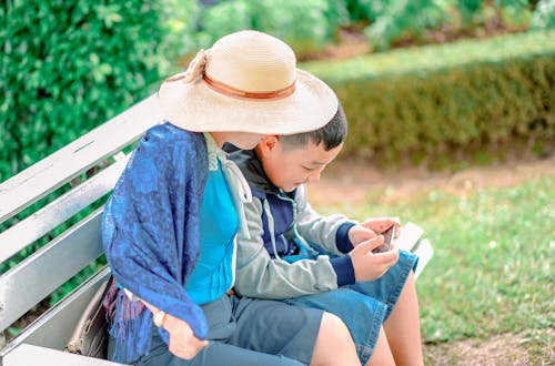男孩身穿灰色，蓝色和黑色拉链夹克，将智能手机紧贴在旁边，身穿米色遮阳帽的人都坐在灰色的木凳上