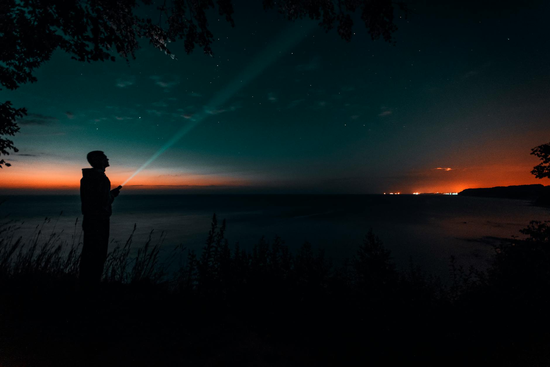 Silhouette of a person with a flashlight by the sea under a starry sky during twilight.