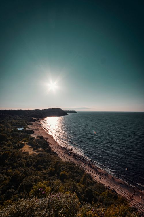Free Serene coastal landscape with bright sun over the ocean and lush greenery. Stock Photo