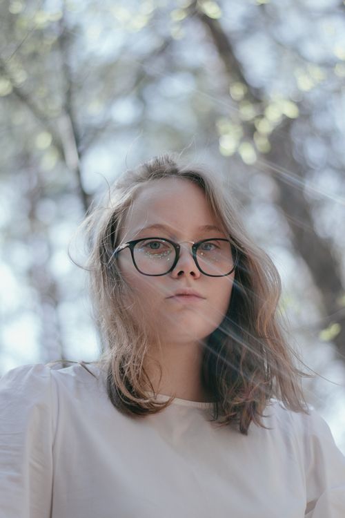 Free Portrait of a young woman with glasses outdoors with sunlight filtering through trees. Stock Photo