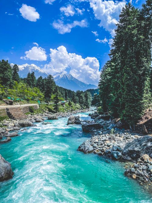 Free Serene view of a river flowing through Pahalgam's lush mountains, framed by a vibrant blue sky and white clouds. Stock Photo