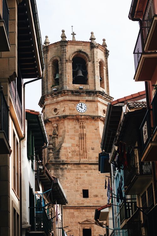 Free A low angle view of a Gothic church tower in Getaria, Spain with a clock face and intricate stonework. Stock Photo