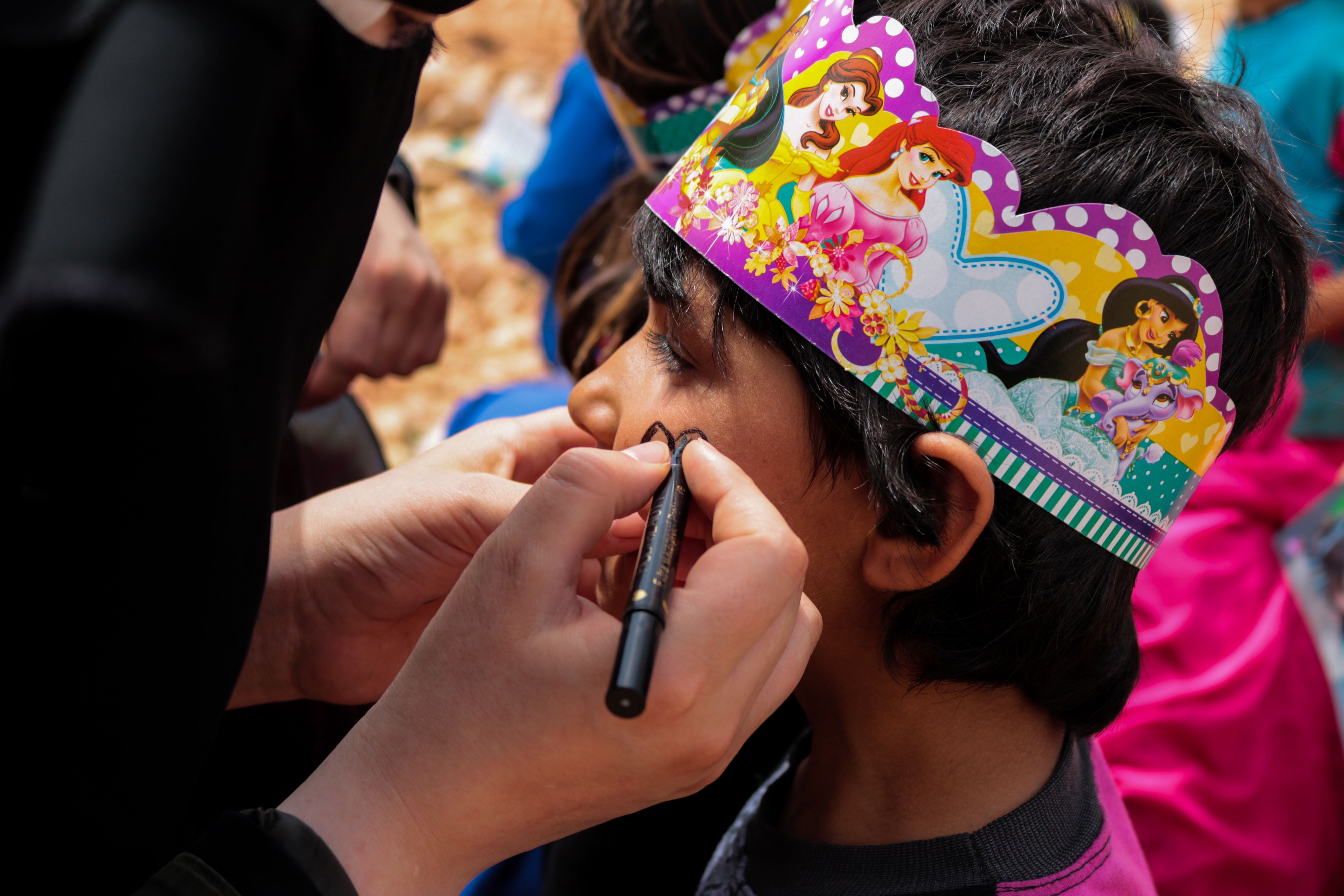 British school end of year party Cairo — A young child in a colorful crown getting face paint applied at an outdoor festival.