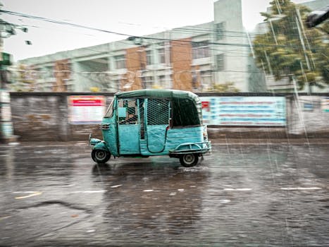 A blue autorickshaw drives through a rainy street in urban Dhaka, Bangladesh.