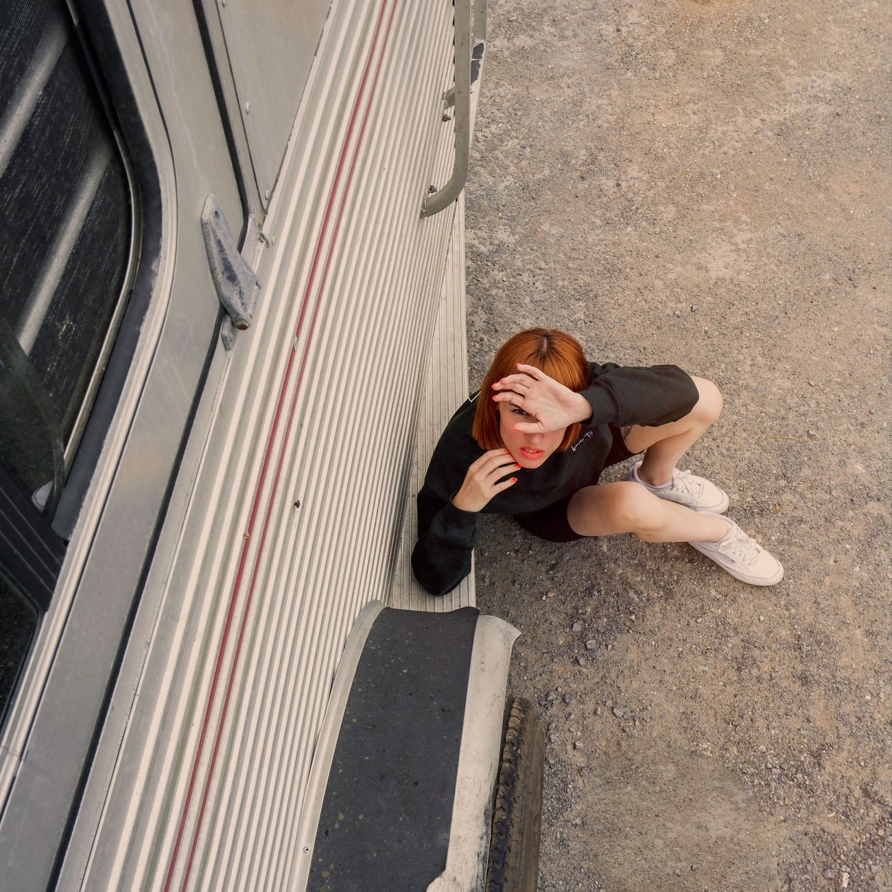 Red-haired woman sitting on dirt ground beside a mobile home, shielding her eyes from the sun.