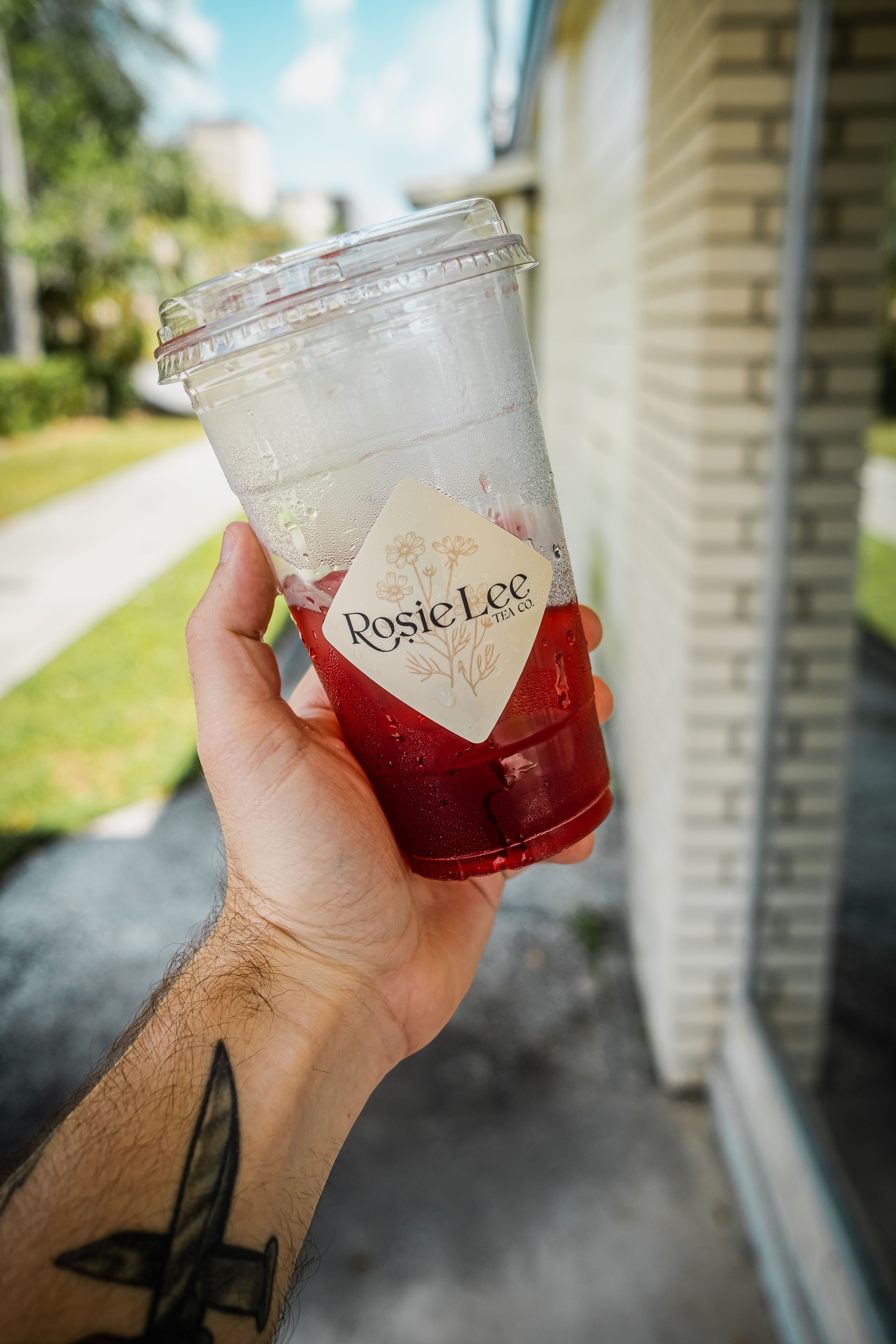 Tattooed hand holding a cold red beverage in clear cup outdoors on a sunny day.