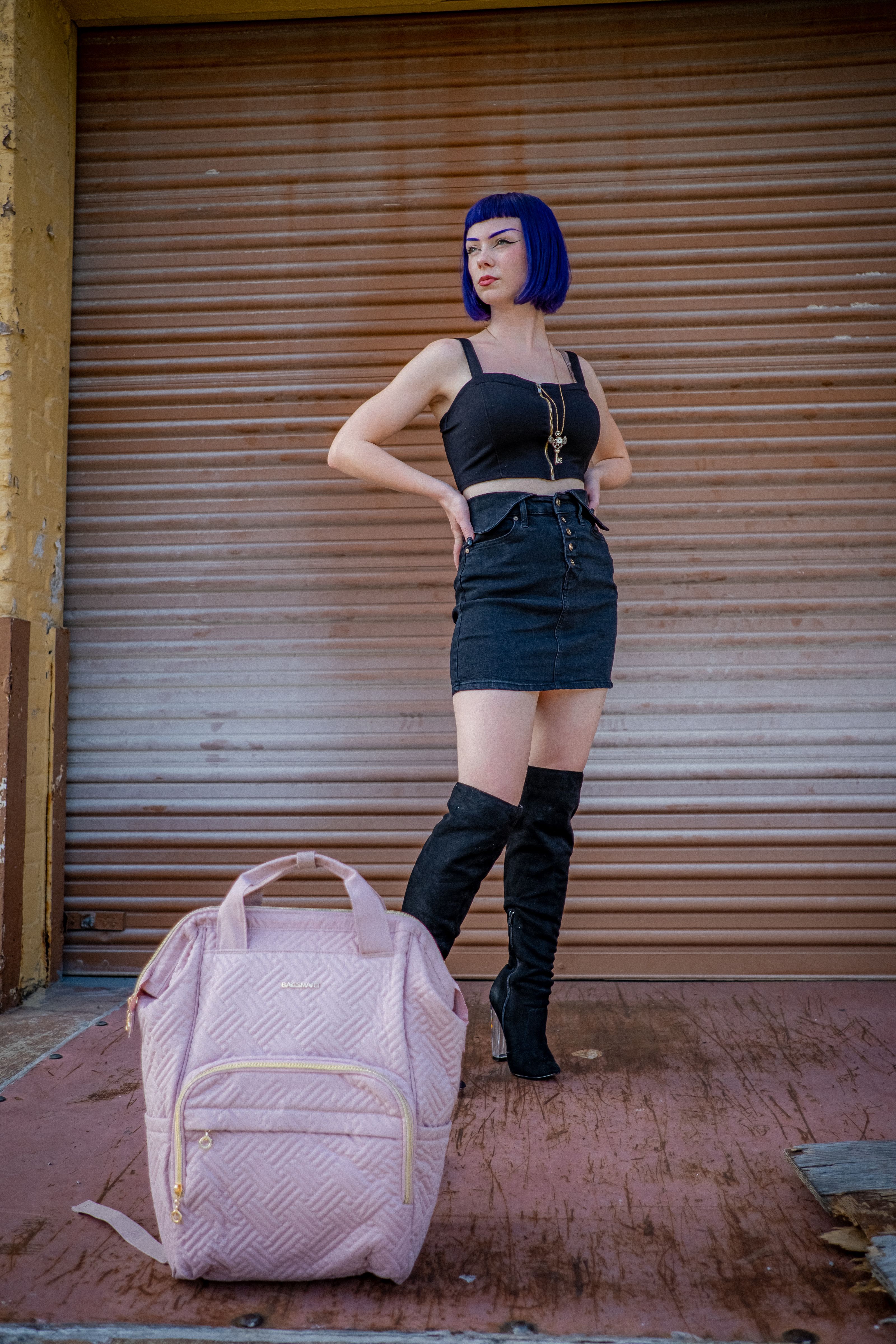 Mulher estilosa com cabelo azul fica confiante com as mãos nos quadris, mochila rosa em primeiro plano.