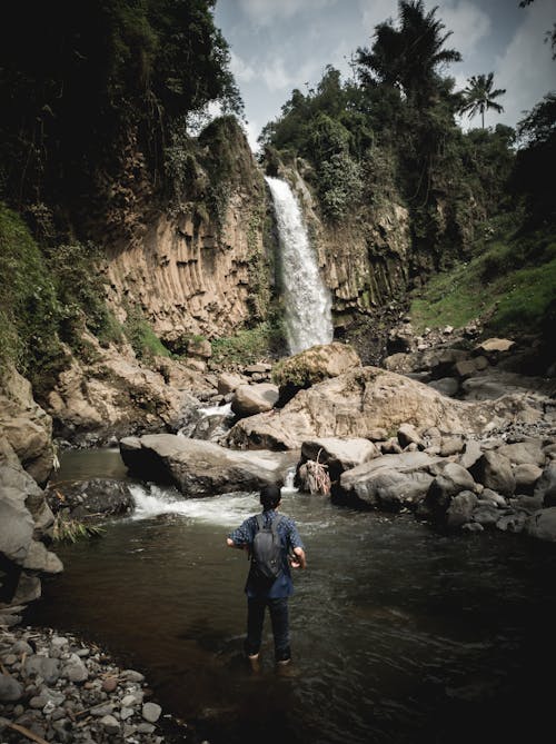 Free Man in Blue Jacket and Black Pants Standing on Brown Rock Near Waterfalls Stock Photo