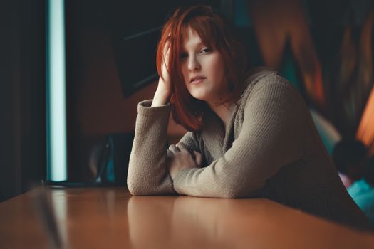 Redhead woman casually sitting at a café table with warm lighting, indoors.