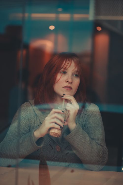 Free Redheaded woman sitting at café window, looking thoughtful with a drink in hand. Stock Photo
