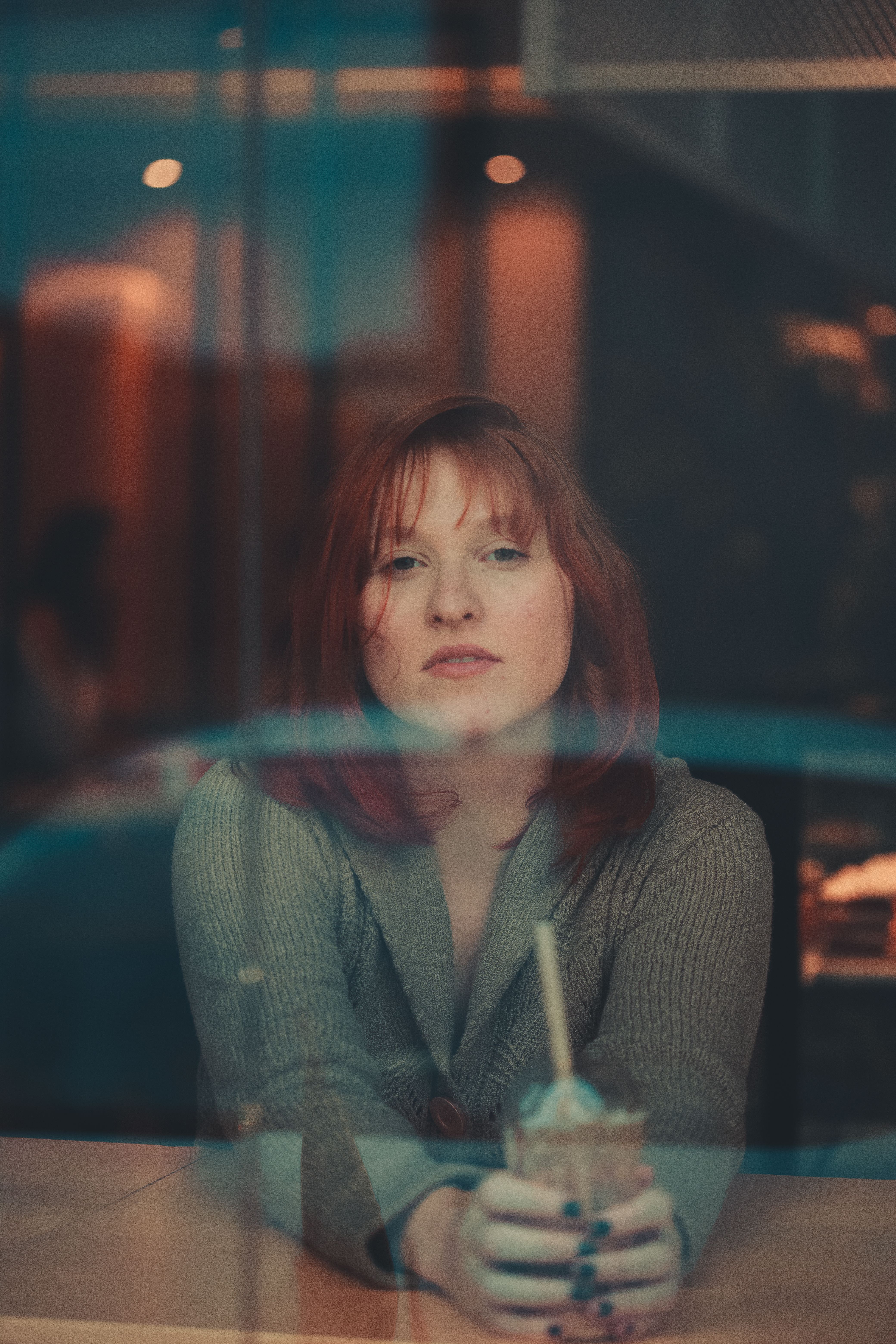 A woman with red hair gazes through a cafe window, holding a milkshake.