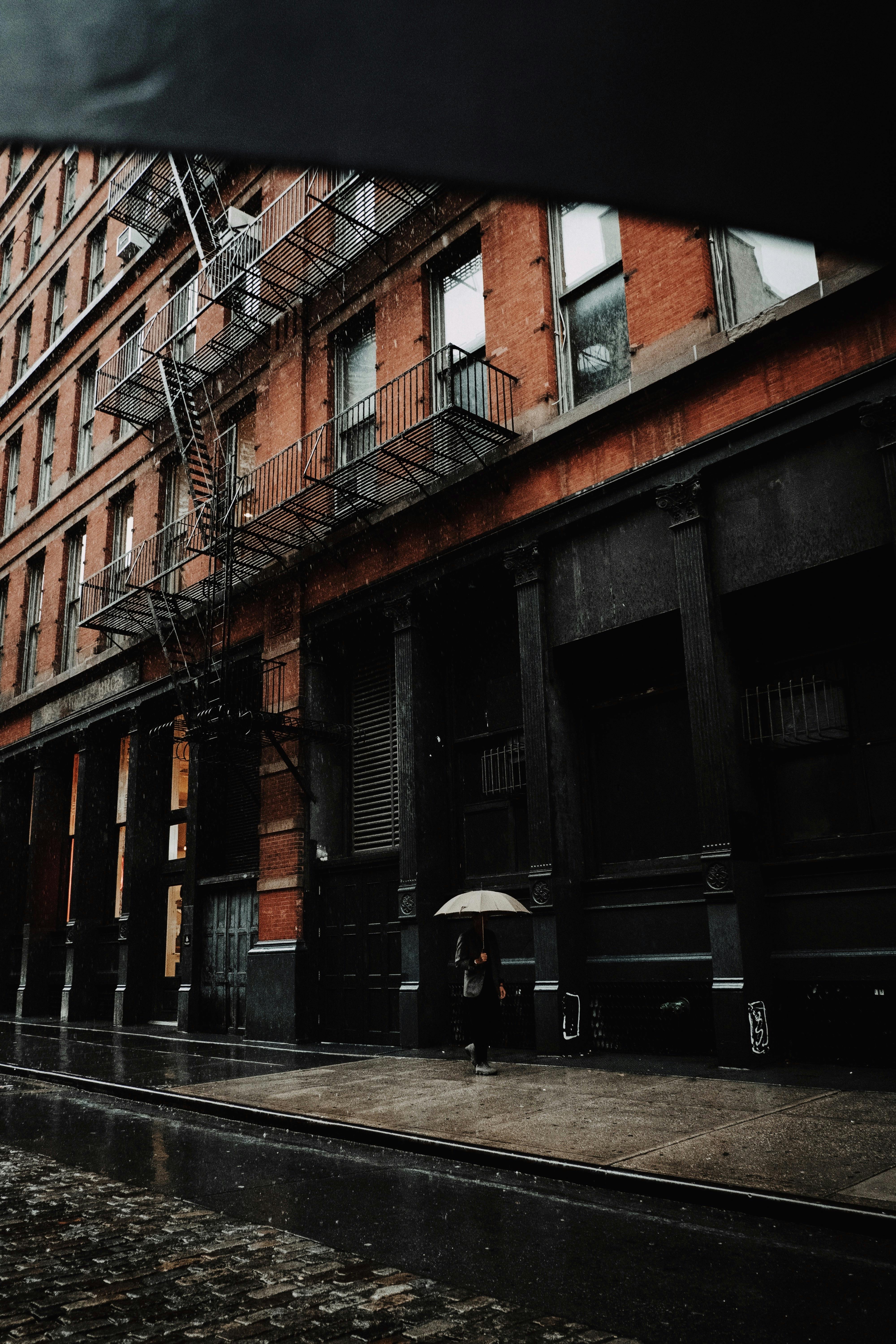 Moody urban street with a person holding an umbrella on a rainy day, featuring a fire escape.