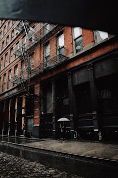 Moody urban street with a person holding an umbrella on a rainy day, featuring a fire escape.