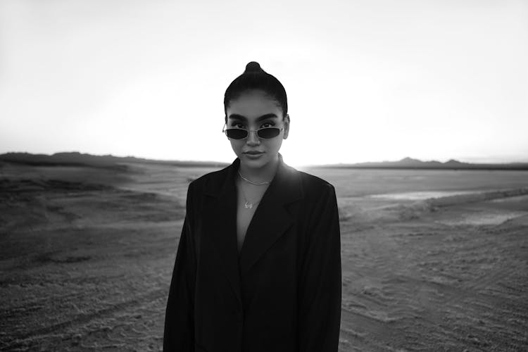 Black And White Portrait Of Woman Wearing Sunglasses And Suit Standing In Empty Field