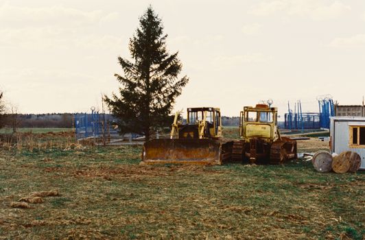 Two bulldozers idle in a rural construction site, surrounded by fields.