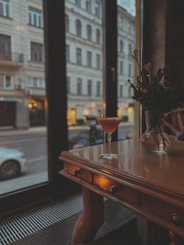 Elegant cocktail on a wooden table by a city window, creating a cozy cafe ambiance.