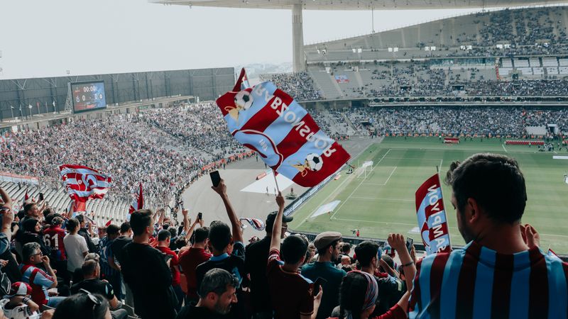 Vibrant crowd cheers at a football match in a large stadium, waving team flags.