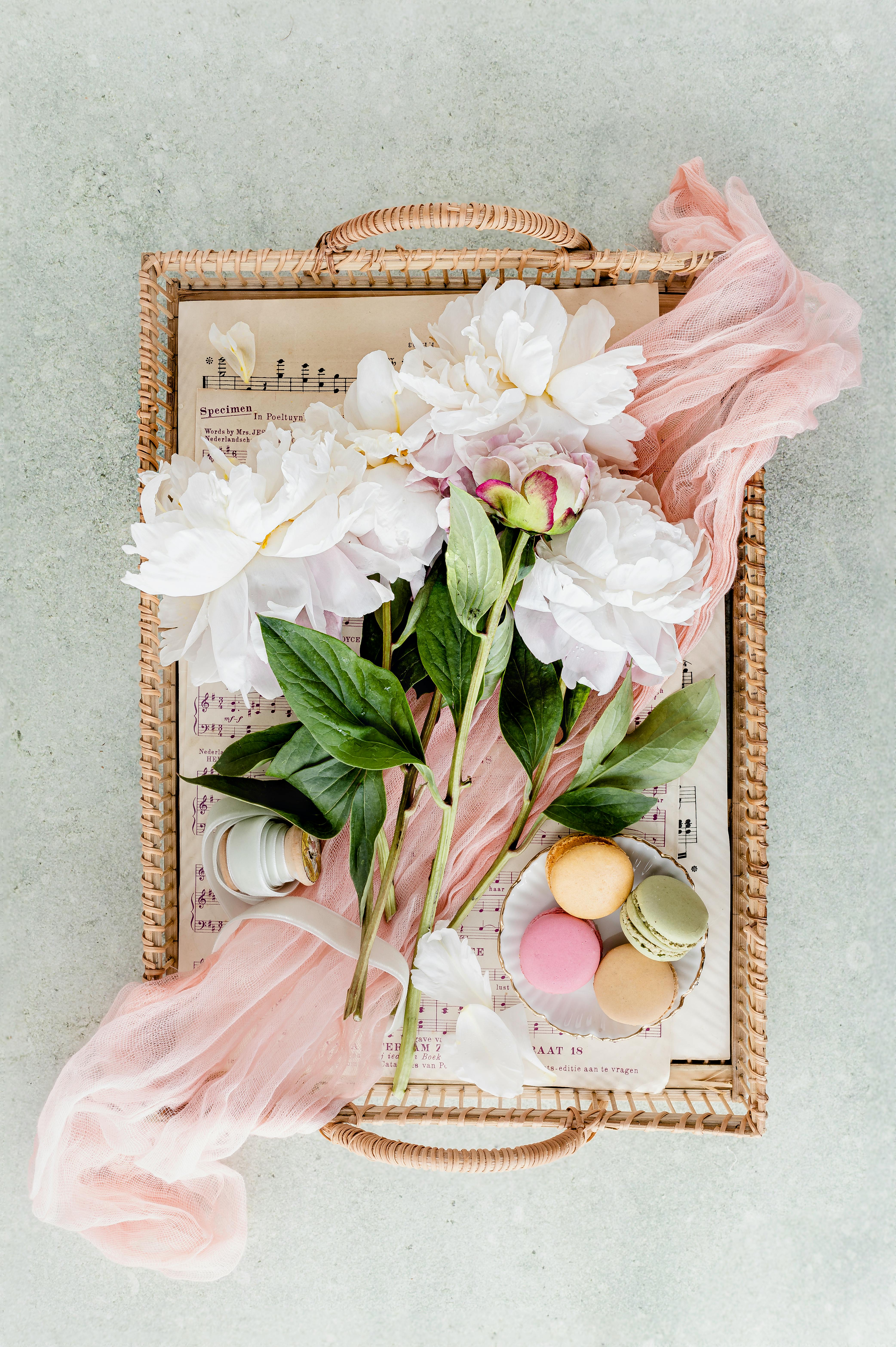Chic flatlay of peonies, macarons, and sheet music on a woven tray for a delicate touch.