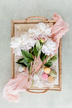 Chic flatlay of peonies, macarons, and sheet music on a woven tray for a delicate touch.