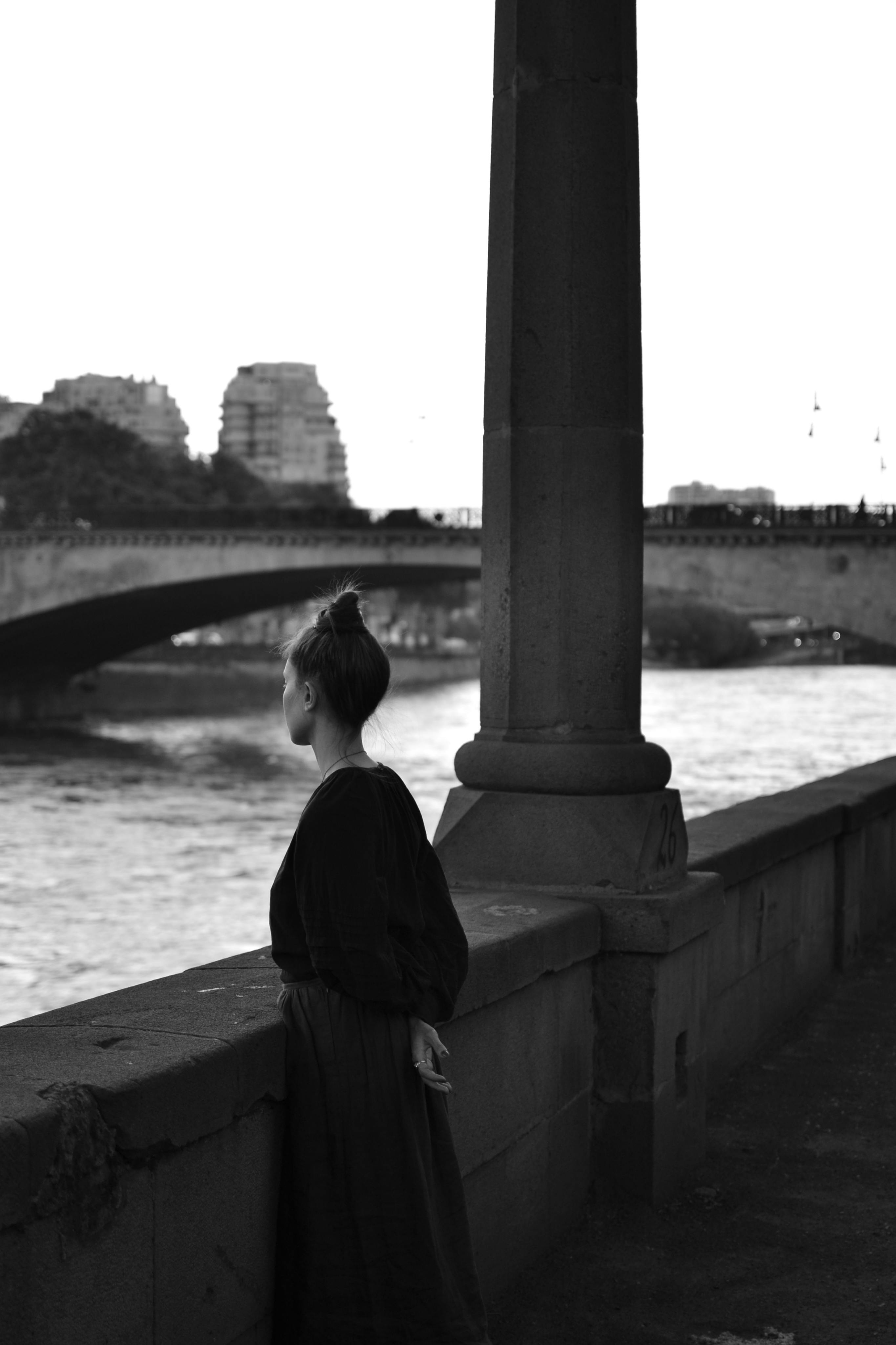 Black and white photo of a woman looking at a bridge over the river.
