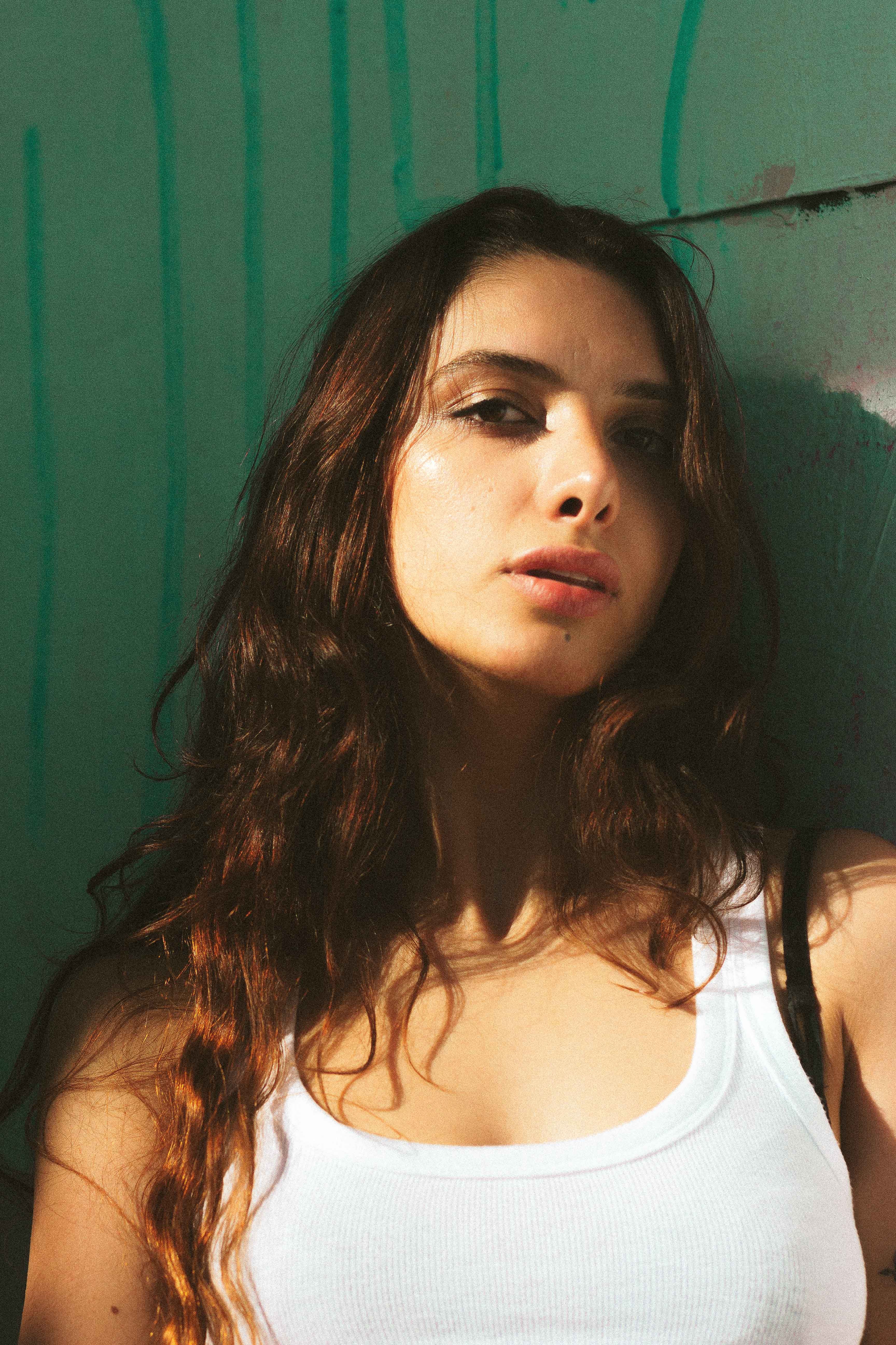 Close-up portrait of a brunette woman wearing a white tank top against a green background.