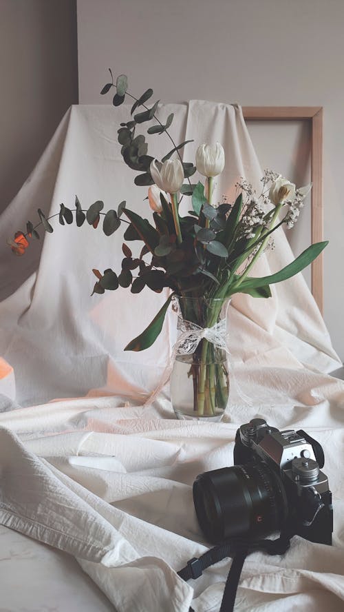 A White Flowers and Green Leaves on a Glass Vase Near the Black Camera