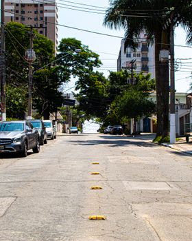 A quiet urban street with parked cars, green trees, and residential buildings on a sunny day.