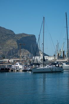 Sailboats docked in a marina with a mountainous backdrop under clear blue skies.
