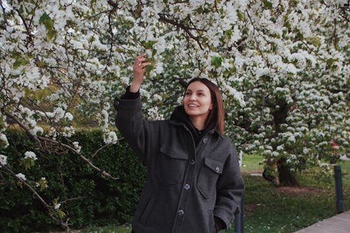 A Woman in Gray Jacket Smiling while Touching White Flowers on the Tree