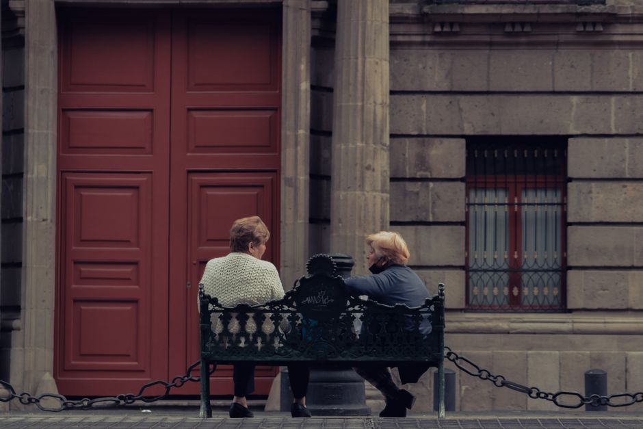 Dos mujeres mayores sentadas y conversando en un banco decorativo fuera de un edificio.