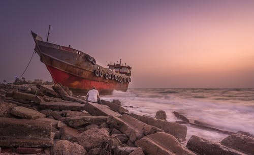 免费 一艘船和一个人坐在海边的石头上的红色蓝色和黑色照片 素材图片