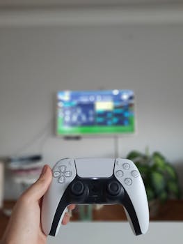 Close-up of hand holding a wireless gaming controller with blurred TV in background.