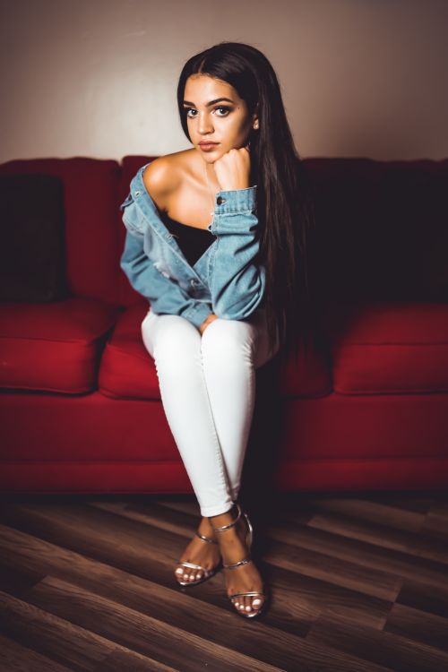 Free Young woman in denim jacket and white pants posing indoors on a red couch. Stock Photo