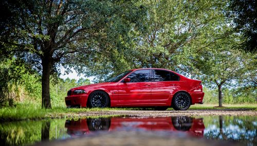 Free A stunning red classic car parked beside a serene lake surrounded by lush greenery. Stock Photo