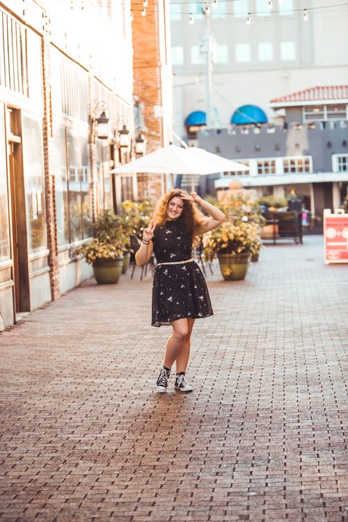 Free Chic young woman in a black dress making a peace sign while smiling on a city sidewalk. Stock Photo