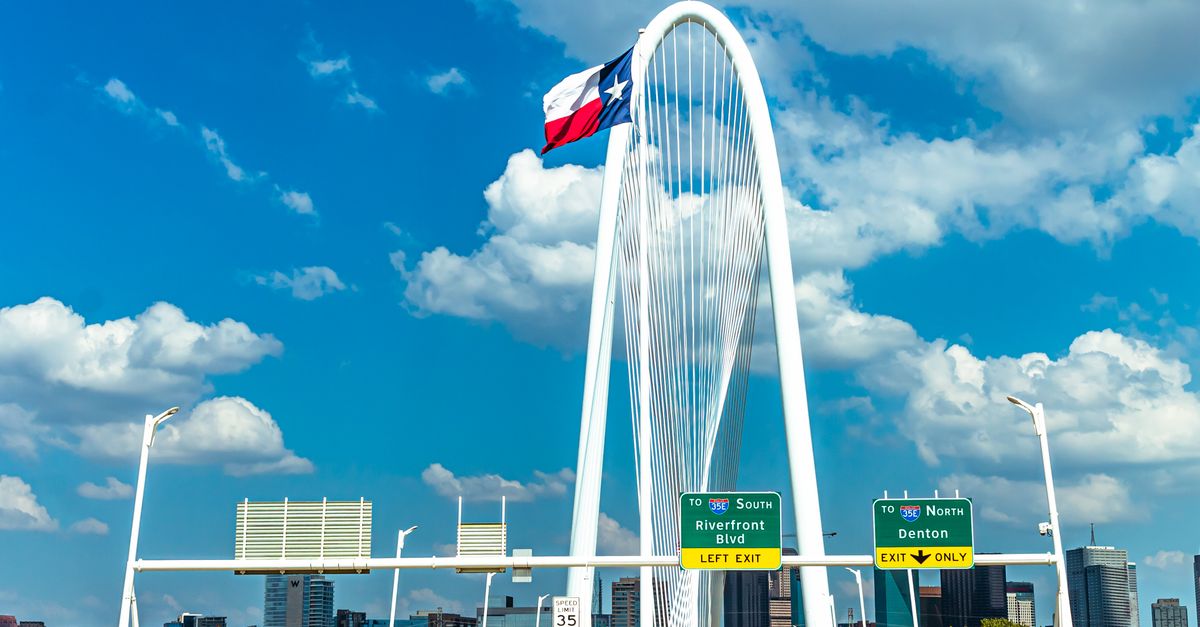 Iconic view of Margaret Hunt Hill Bridge with Texas flag and Dallas skyline under a blue sky.