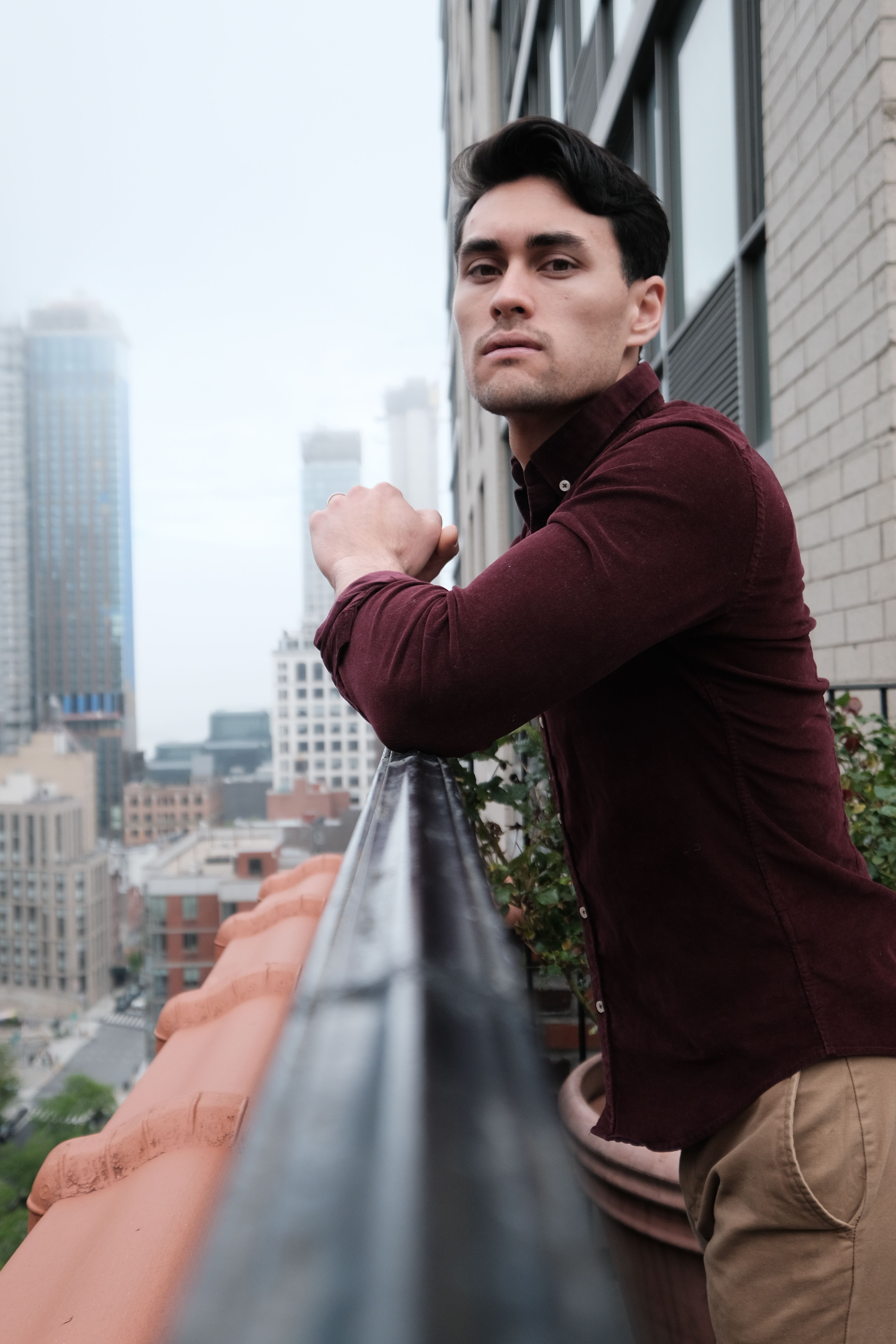 A man with black hair leans on a railing with a city skyline in the background.