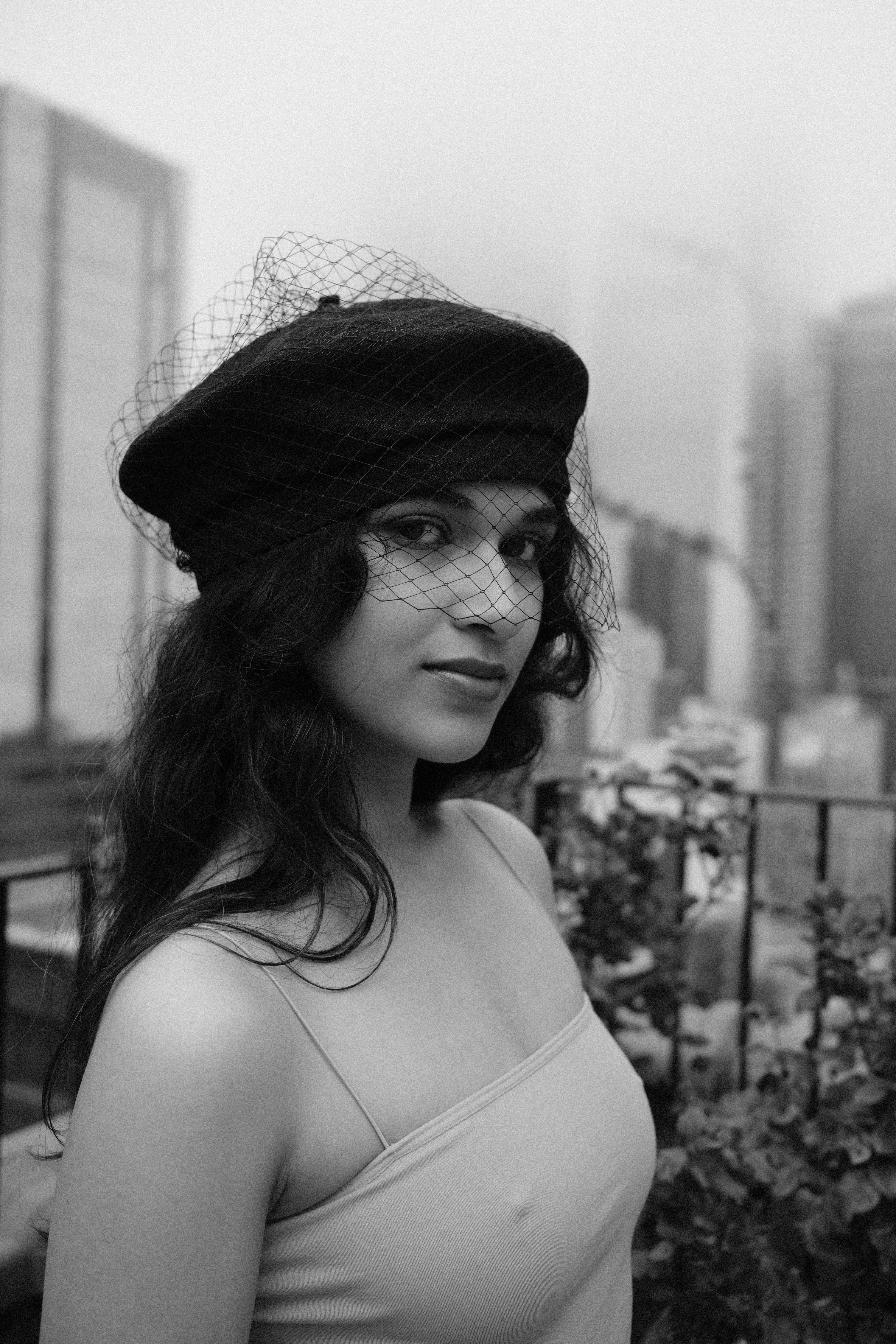 A stylish woman poses with a beret and veil in a black and white cityscape portrait.