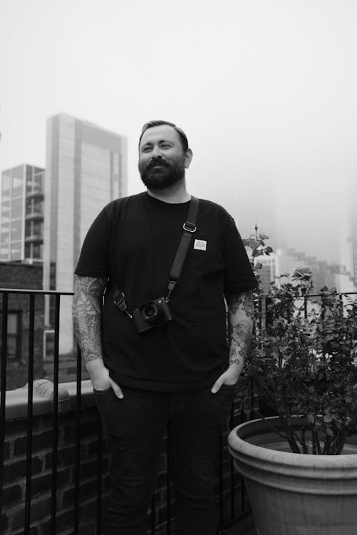 A Grayscale Photo of a Tattooed Man in Black Shirt Standing Near the Metal Railings