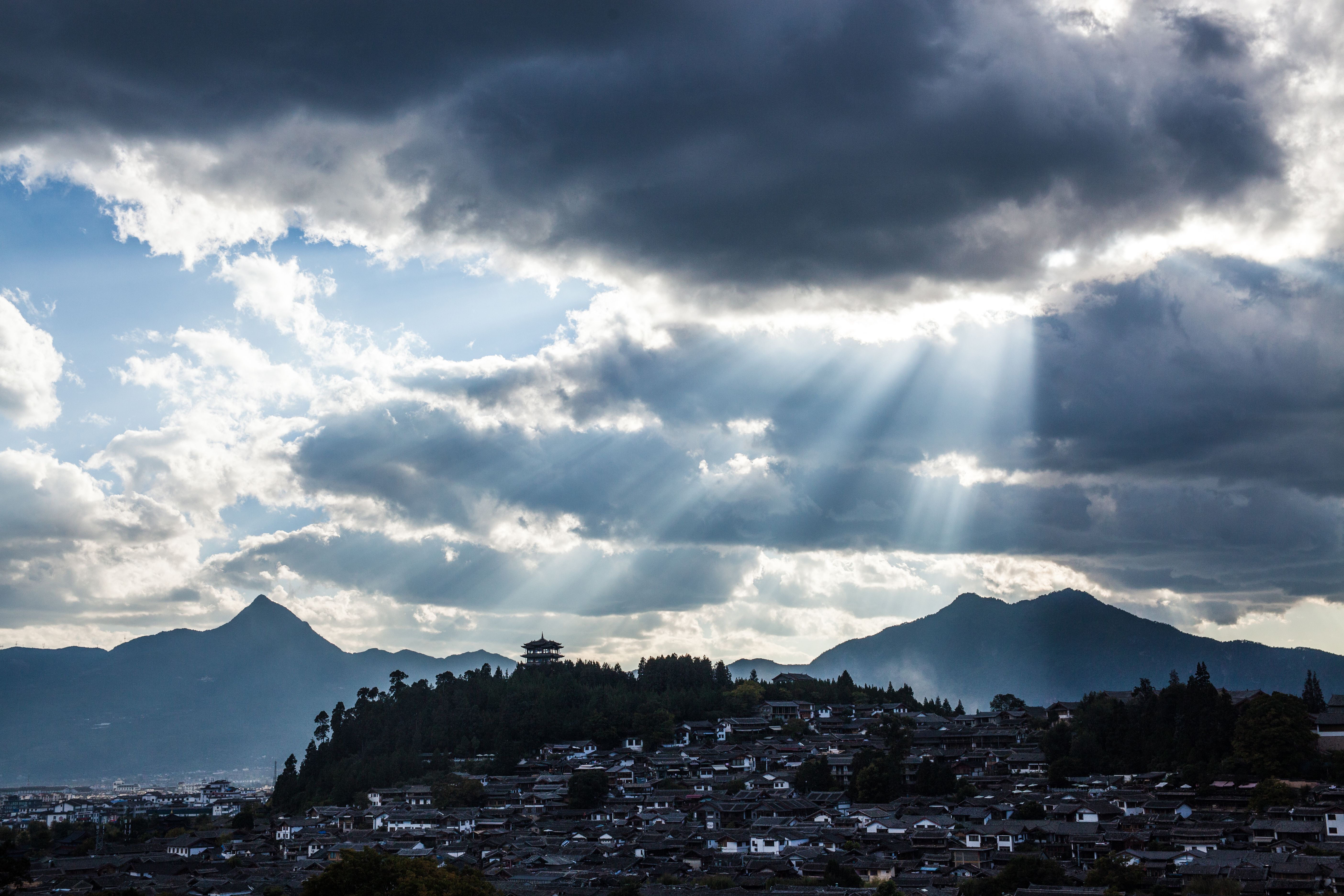 A dramatic cityscape with cloud-filled skies and sunbeams breaking through mountainous terrain.