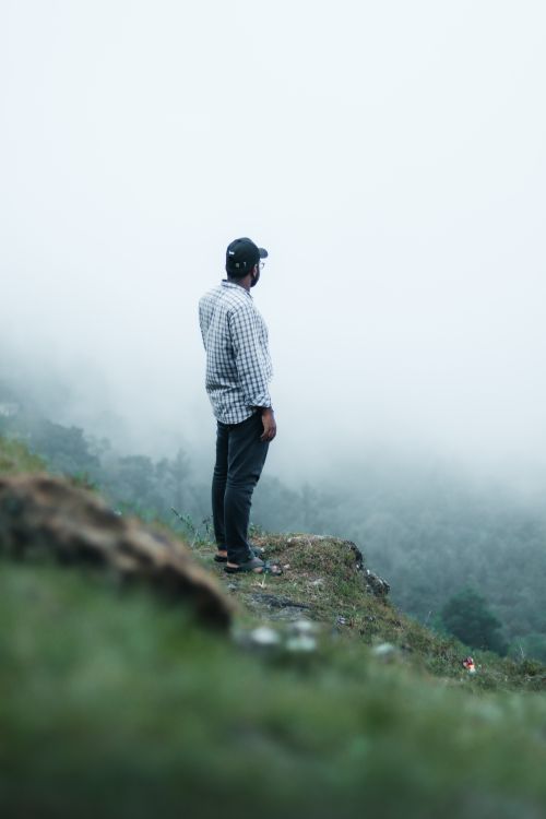 Free Man wearing a cap gazing into foggy landscape from a cliff in KL, India. Stock Photo